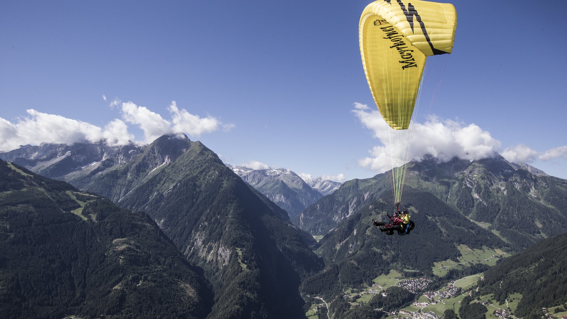 Unforgettable paragliding experience Paragliders over green mountains with a wide valley and blue sky in the background.
