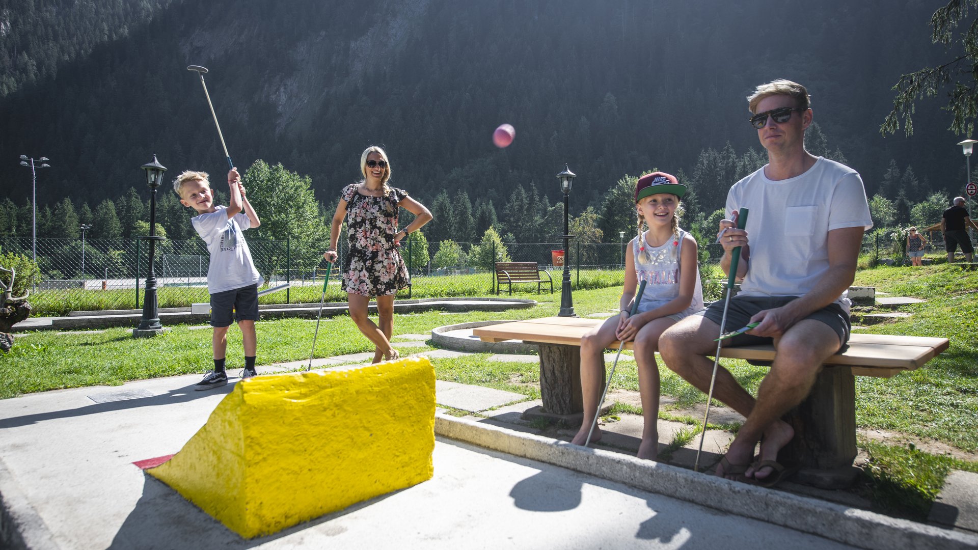 Family minigolf adventure Family playing mini golf outdoors, surrounded by green mountains and trees.