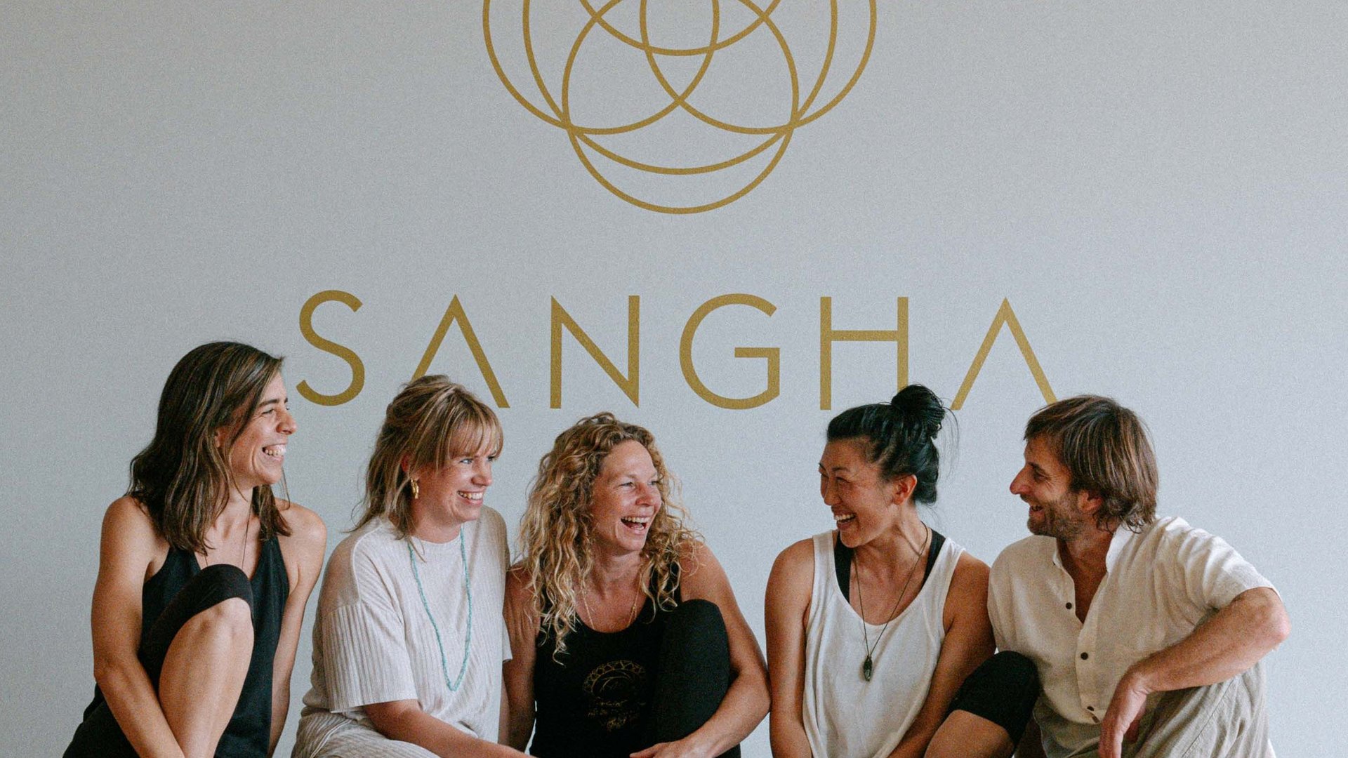Five people sit smiling on the floor in front of a wall with the "SANGHA" logo.